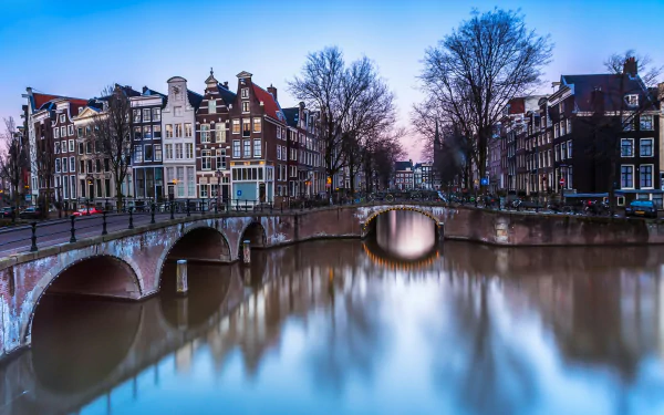 4K Ultra HD image of Amsterdam’s iconic canal with historic buildings and stone bridges, showcasing man-made city architecture at dusk.