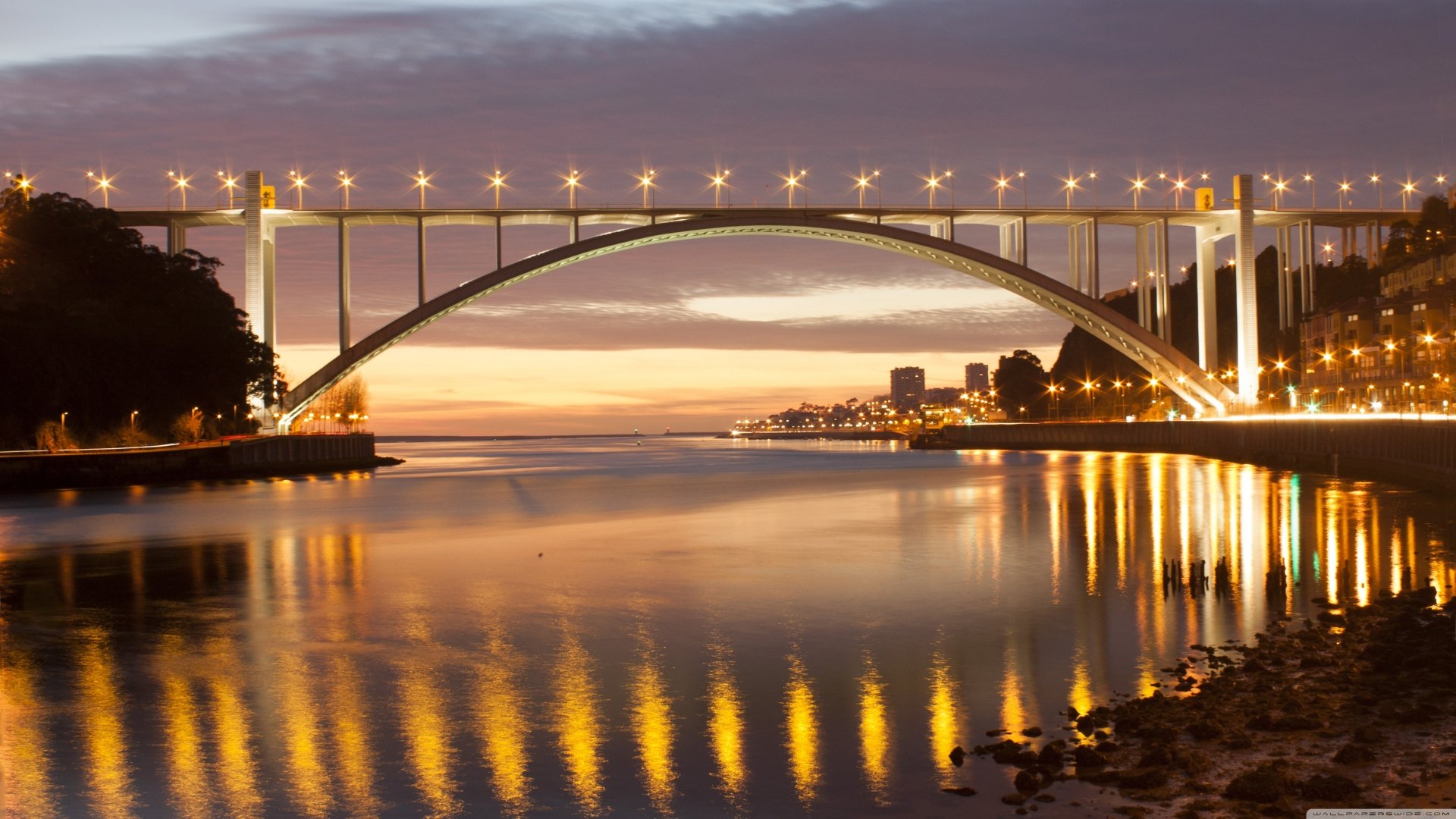 4K Ultra HD wallpaper of Portugal's man-made Arrábida Bridge illuminated at dusk, reflecting on calm waters with city lights in the background.