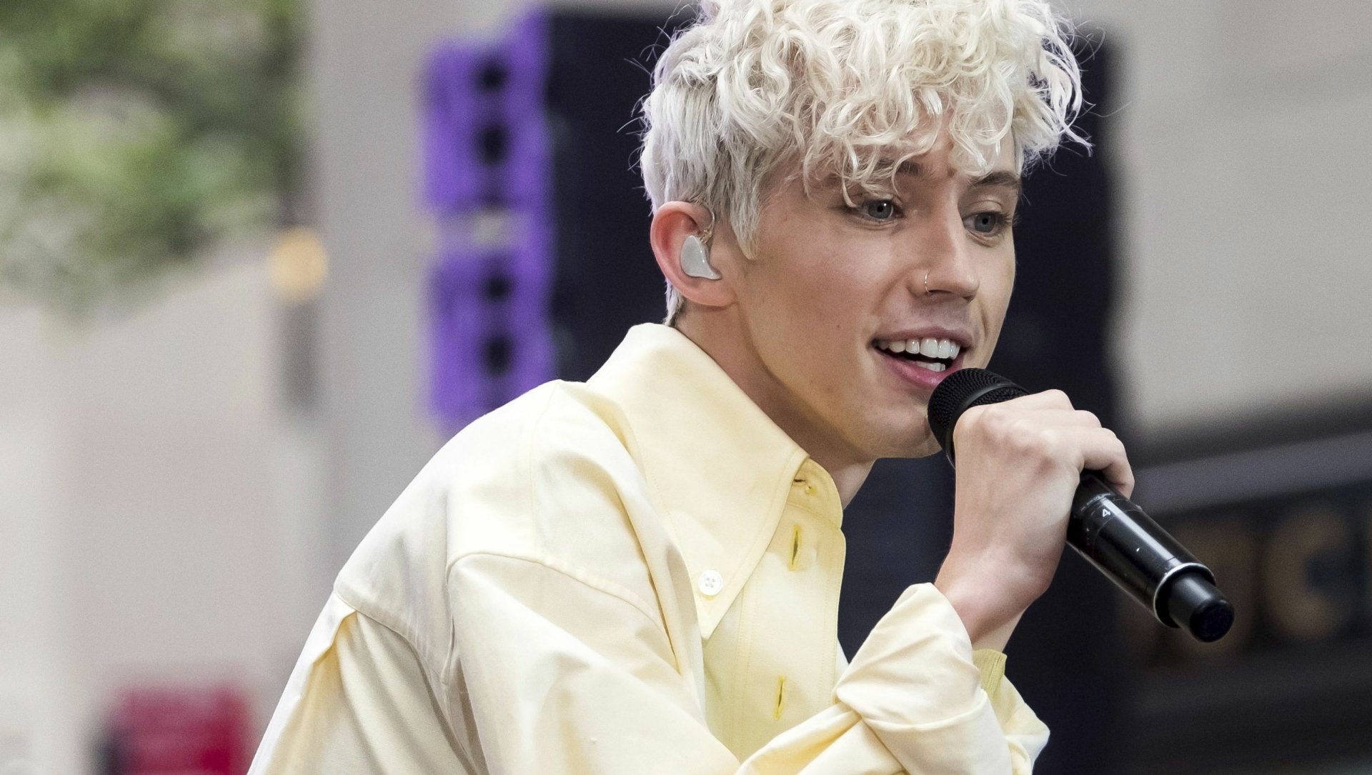 HD desktop wallpaper featuring a person singing into a microphone, wearing a pale yellow shirt, with a blurred background.