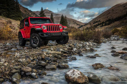 Red Jeep Wrangler SUV parked on rocky terrain beside a flowing stream, surrounded by mountains and trees under a cloudy sky in this HD PC desktop wallpaper.