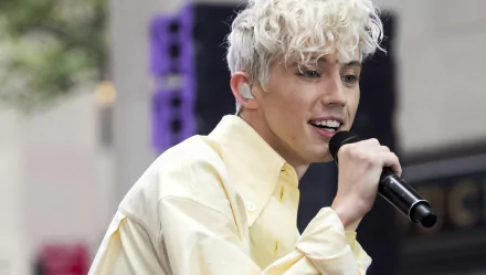 HD desktop wallpaper featuring a person singing into a microphone, wearing a pale yellow shirt, with a blurred background.