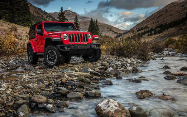Red Jeep Wrangler SUV parked on rocky terrain beside a flowing stream, surrounded by mountains and trees under a cloudy sky in this HD PC desktop wallpaper.