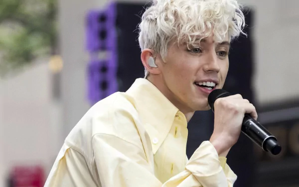 HD desktop wallpaper featuring a person singing into a microphone, wearing a pale yellow shirt, with a blurred background.