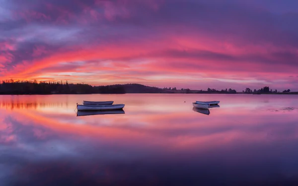 A serene 4K Ultra HD view of Loch Lomond, Scotland at sunset, featuring two boats floating on the calm lake under a vibrant pink and purple sky.