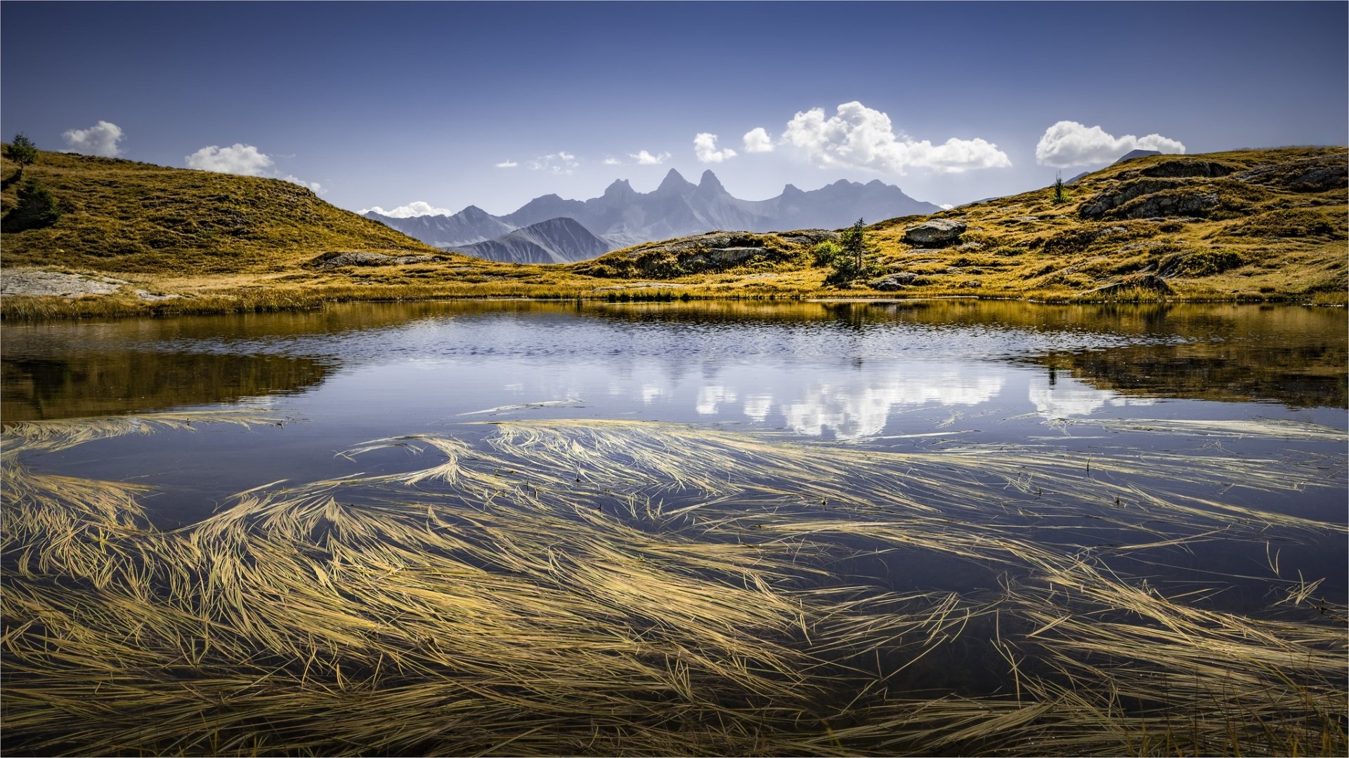 HD PC desktop wallpaper featuring a serene mountain landscape with a clear lake reflecting the sky and distant peaks, surrounded by grassy hills.