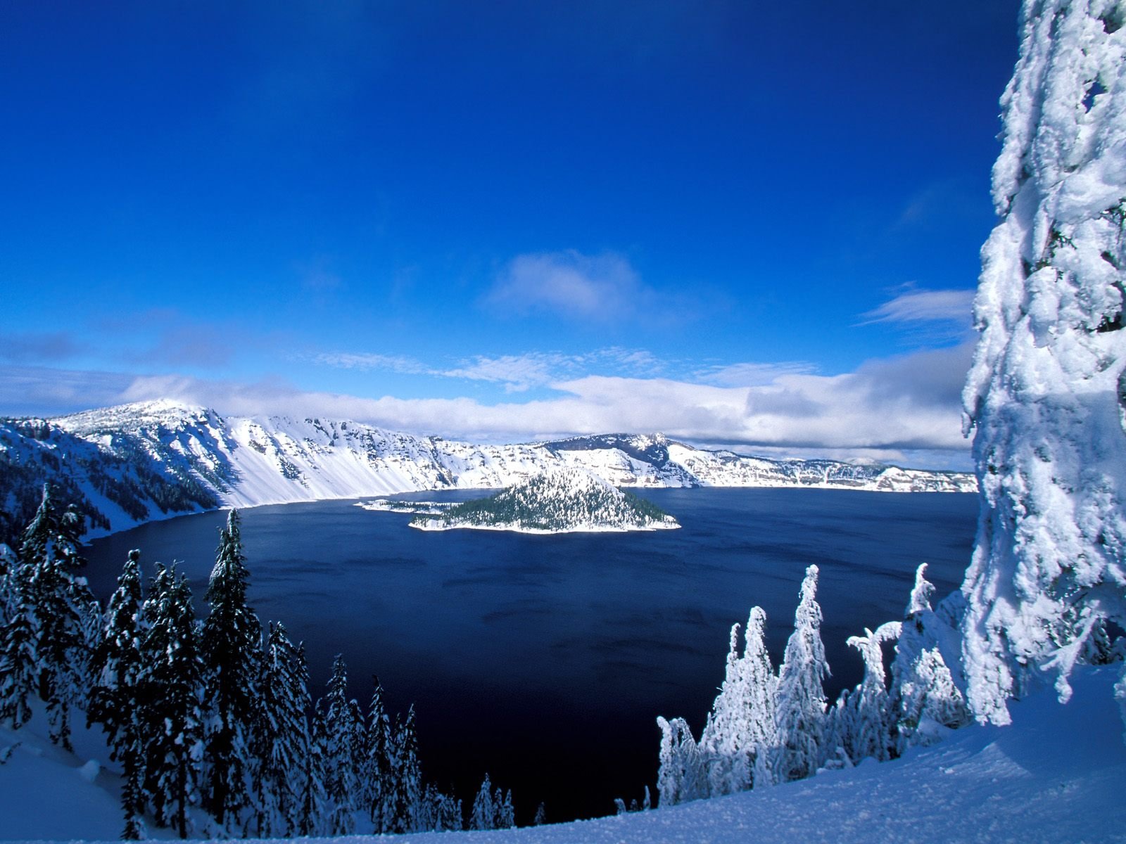 HD PC desktop wallpaper showcasing a serene winter landscape with snow-covered trees framing a deep blue lake beneath a clear sky.