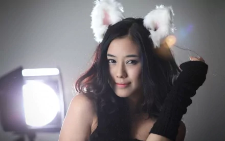 HD desktop wallpaper featuring a pretty woman with long dark hair, wearing black gloves and white fluffy cat ears, posed in front of a soft light background.