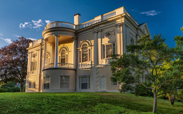HD desktop wallpaper featuring a man-made mansion bathed in warm sunlight, surrounded by green trees and grass under a clear blue sky.