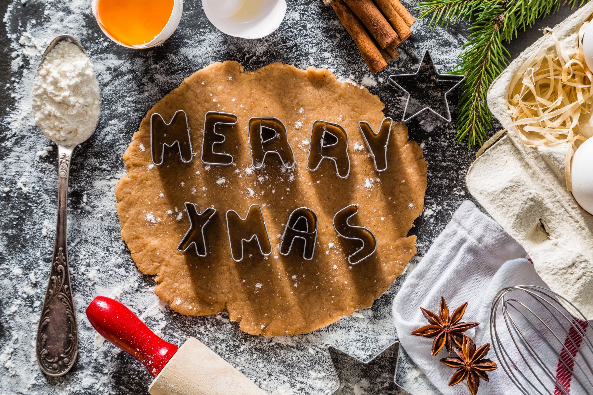 Holiday baking scene: rolled dough stamped MERRY XMAS with flour, rolling pin, spices and cookie cutters — Merry Christmas holiday baking 5K Ultra HD PC desktop wallpaper/background.