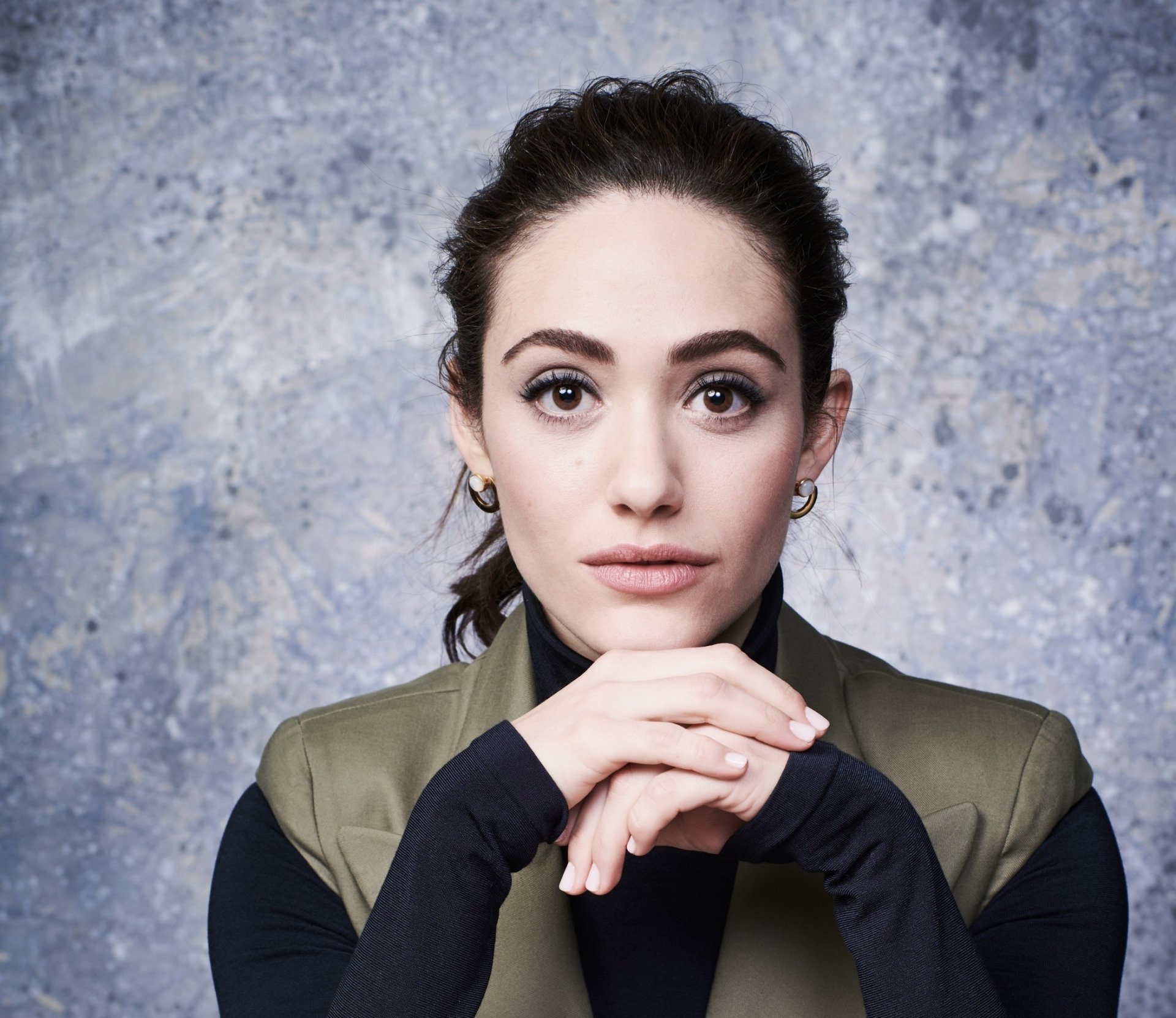 HD desktop wallpaper of a brunette American actress and celebrity with brown eyes, poised against a textured gray background, hands clasped under her chin and looking directly at the camera.