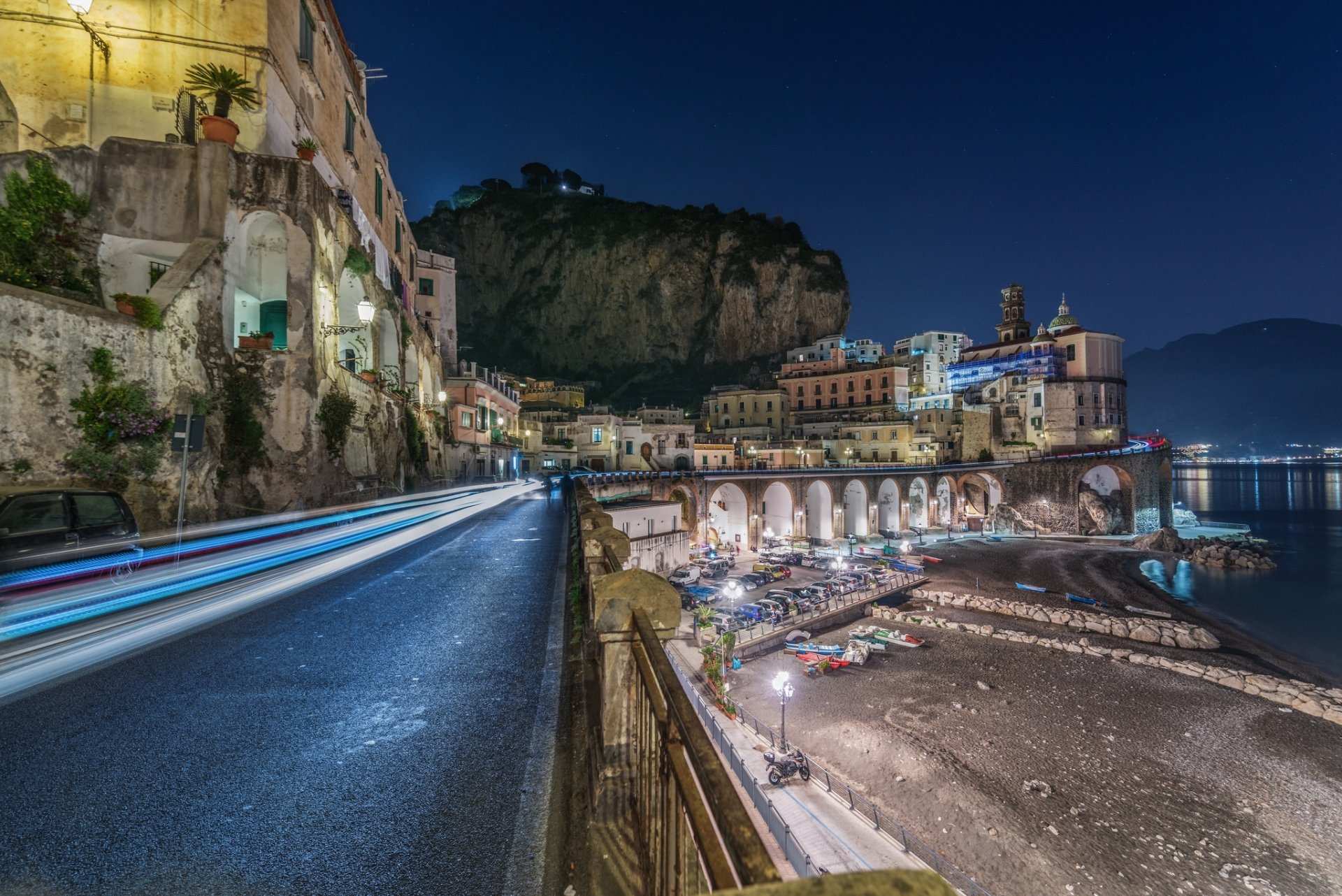 Night view of Amalfi town, Italy — time-lapse long exposure of a coastal road with light trails, arched man-made waterfront buildings and beach. HD PC desktop wallpaper.