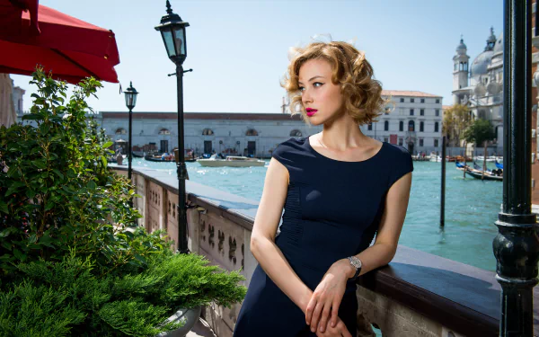 Canadian actress Sarah Gadon with blonde hair, blue eyes, and lipstick wears a blue dress, posing outdoors near water with historic buildings in the background.