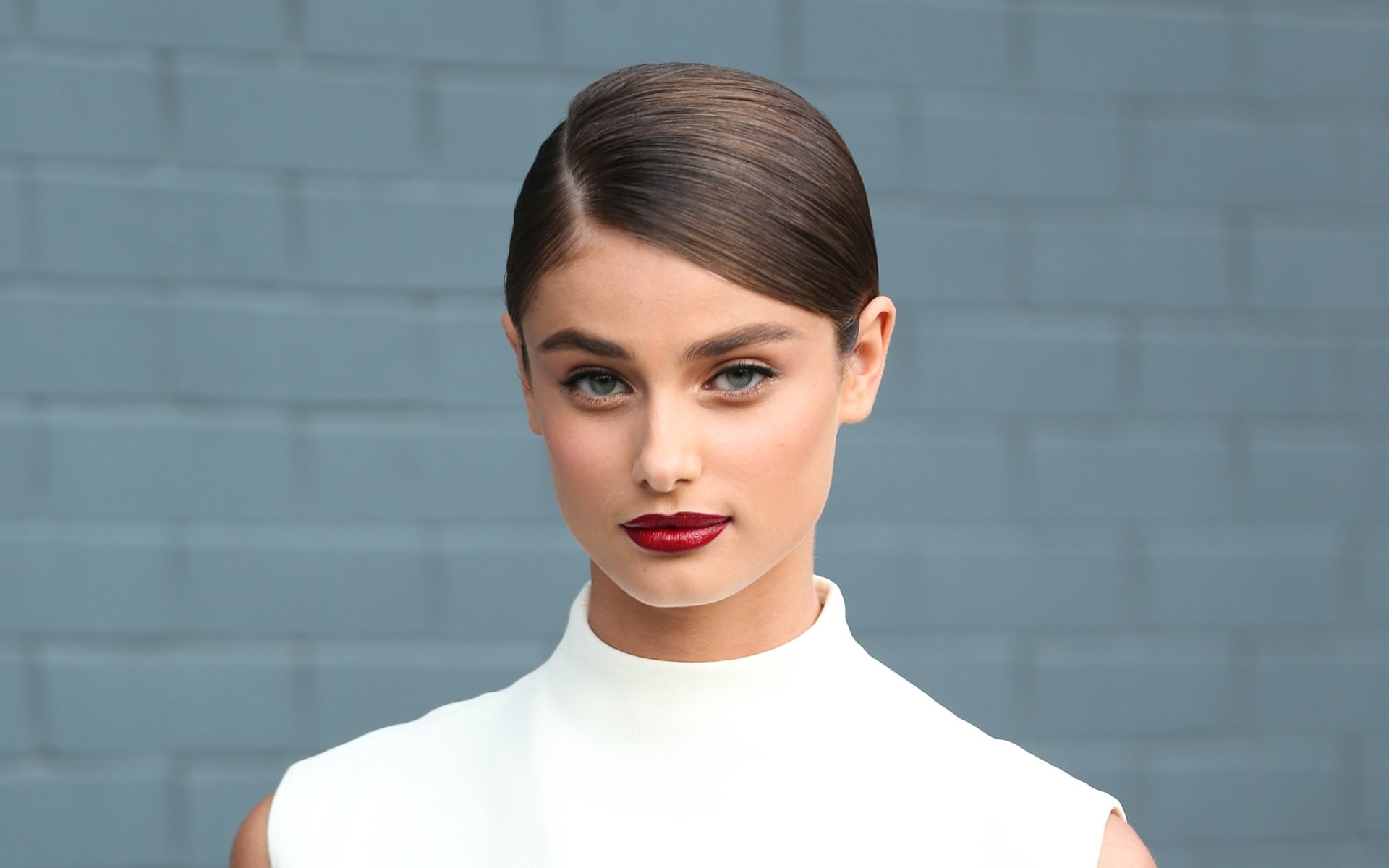 HD desktop wallpaper featuring American brunette model Taylor Marie Hill with sleek hair and bold red lipstick against a muted blue brick wall background.