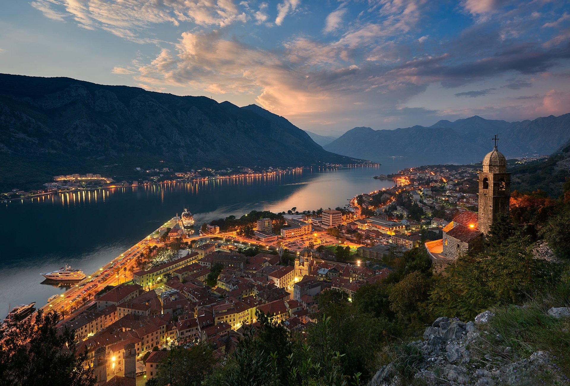 HD desktop wallpaper of a Montenegro town at dusk, featuring a river winding through the man-made landscape with mountains in the background under a colorful sky.
