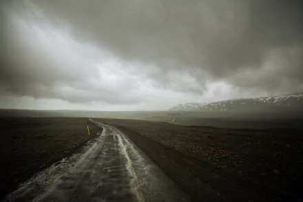 A winding man-made road stretches through a dark Icelandic landscape under heavy clouds, with distant mountains barely visible in this 4K Ultra HD wallpaper.