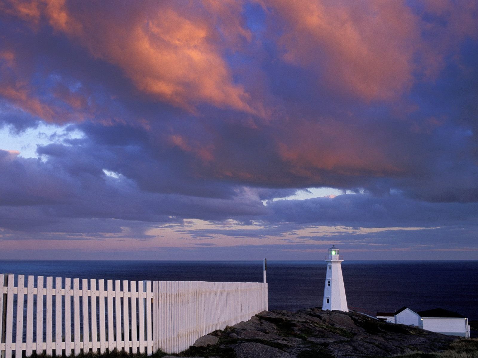 HD PC desktop wallpaper: Newfoundland, Canada coastline at dusk with a man-made white lighthouse on rocky cliffs beneath dramatic purple-orange clouds.