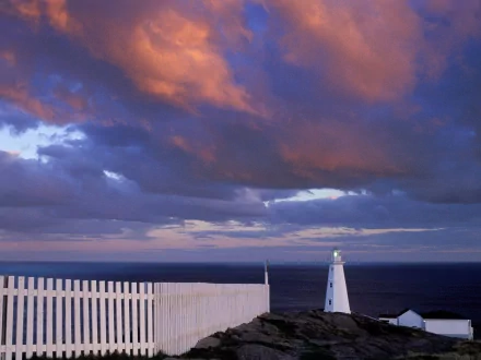 HD PC desktop wallpaper: Newfoundland, Canada coastline at dusk with a man-made white lighthouse on rocky cliffs beneath dramatic purple-orange clouds.