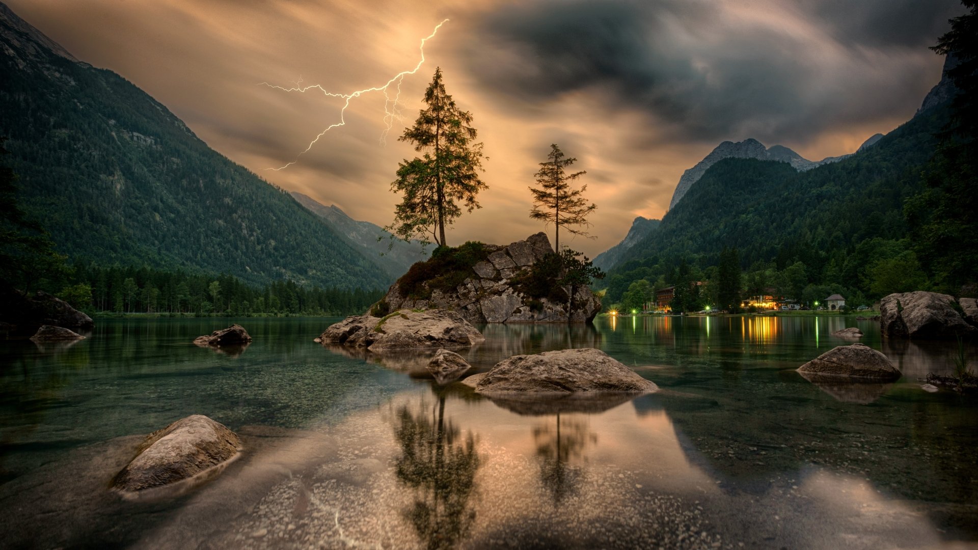 A stunning HD wallpaper featuring a serene island in a tranquil lake, framed by towering mountains. The dramatic sky is illuminated by a lightning strike, adding to the breathtaking natural scenery.