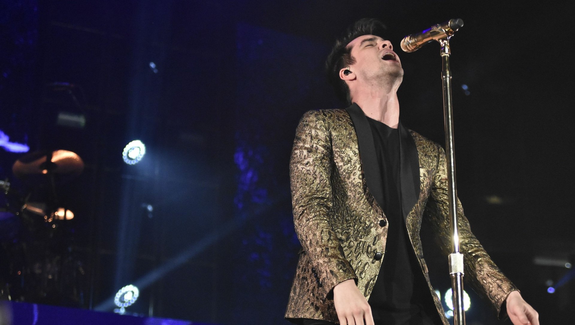 HD wallpaper of a Panic! at the Disco performance with the lead singer on stage in a golden jacket, singing into a microphone.